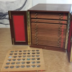 A wooden storage cabinet with one tray removed. Coins are placed in each of the round depressions.