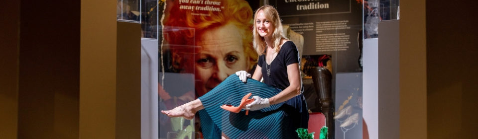 A woman sat on a chair wearing, white gloves and holding an orange, high-heeled shoe.