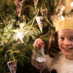 A girl wearing a Christmas cracker hat holding a Christmas tree decoration.