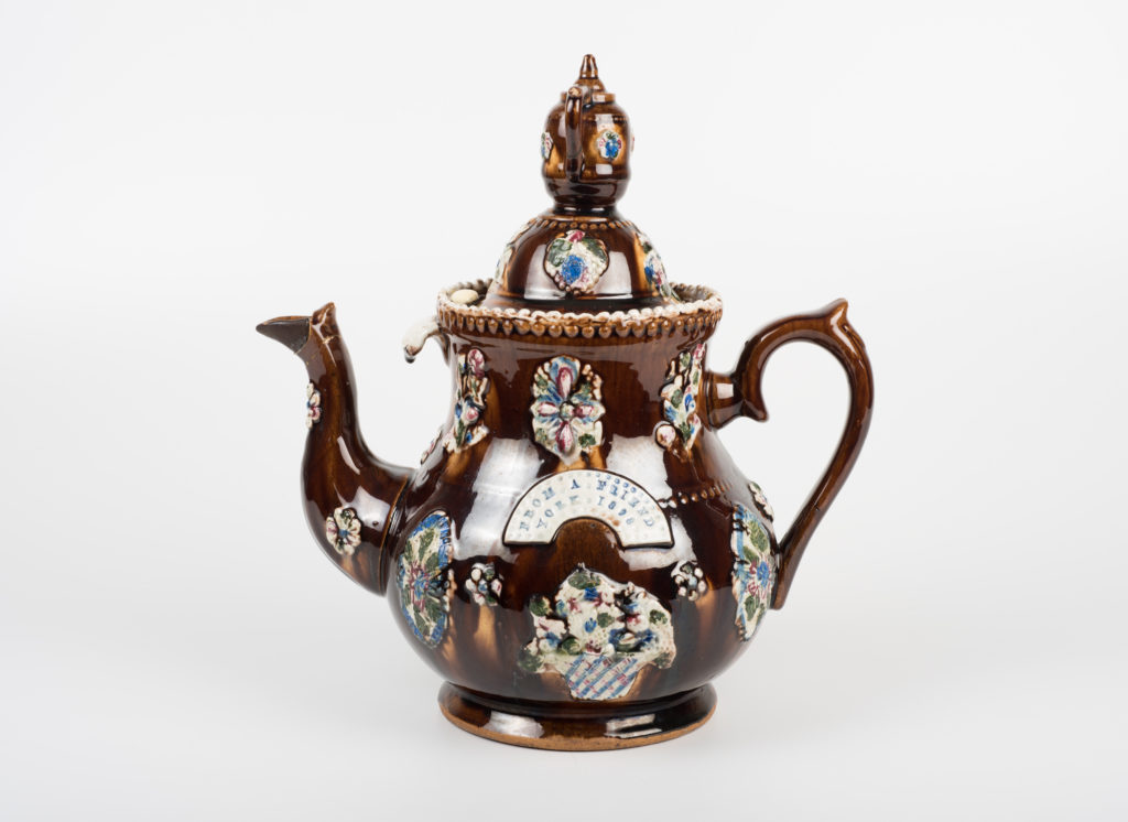 A brown ornamental teapot decorated with badges, along with a label which says 'from a friend, York [illegible date]'