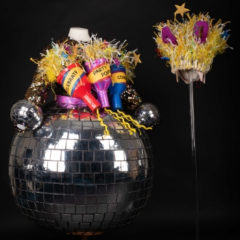 A colourful sequin dress with three giant party poppers stuck to it, and a piece of headgear made of various coloured ribbons and stars.