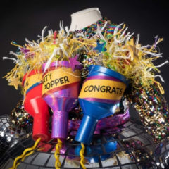 A colourful sequin dress with three giant party poppers stuck to it, one which says 'congrats'.
