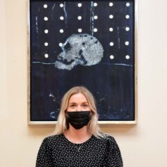 Woman stands in front of an artwork, she wears a black dress and a black mask. The artwork is a Black painting with a skull in the middle. The upper half of the work has a series of white dots. The work is framed in a silver border.